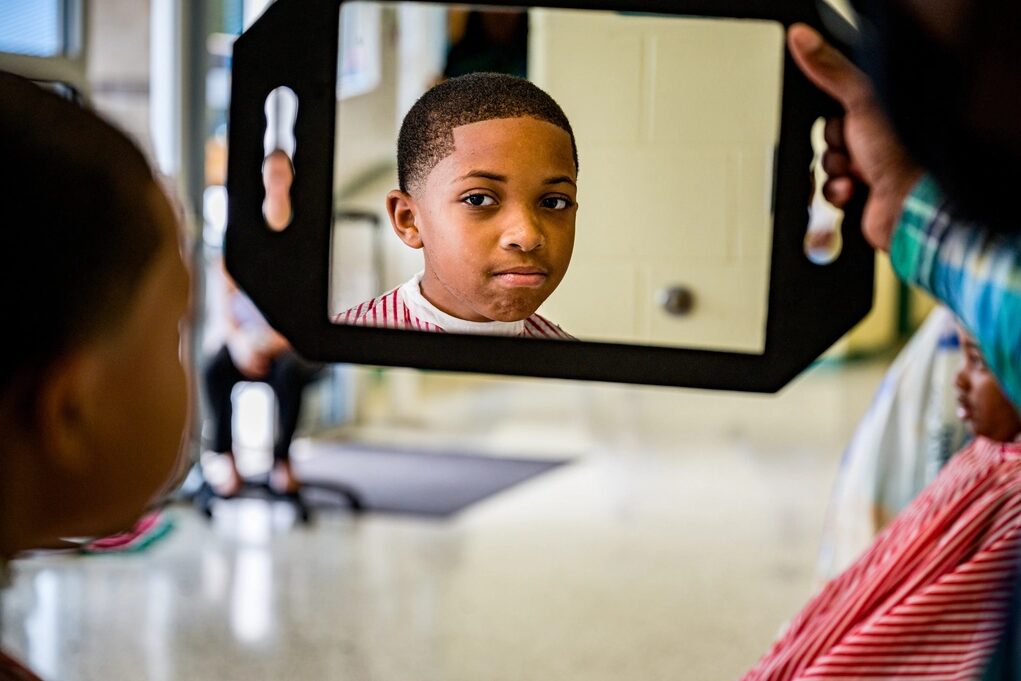 Niño con su corte recién hecho mirándose en el espejo de mano que le sostiene el barbero